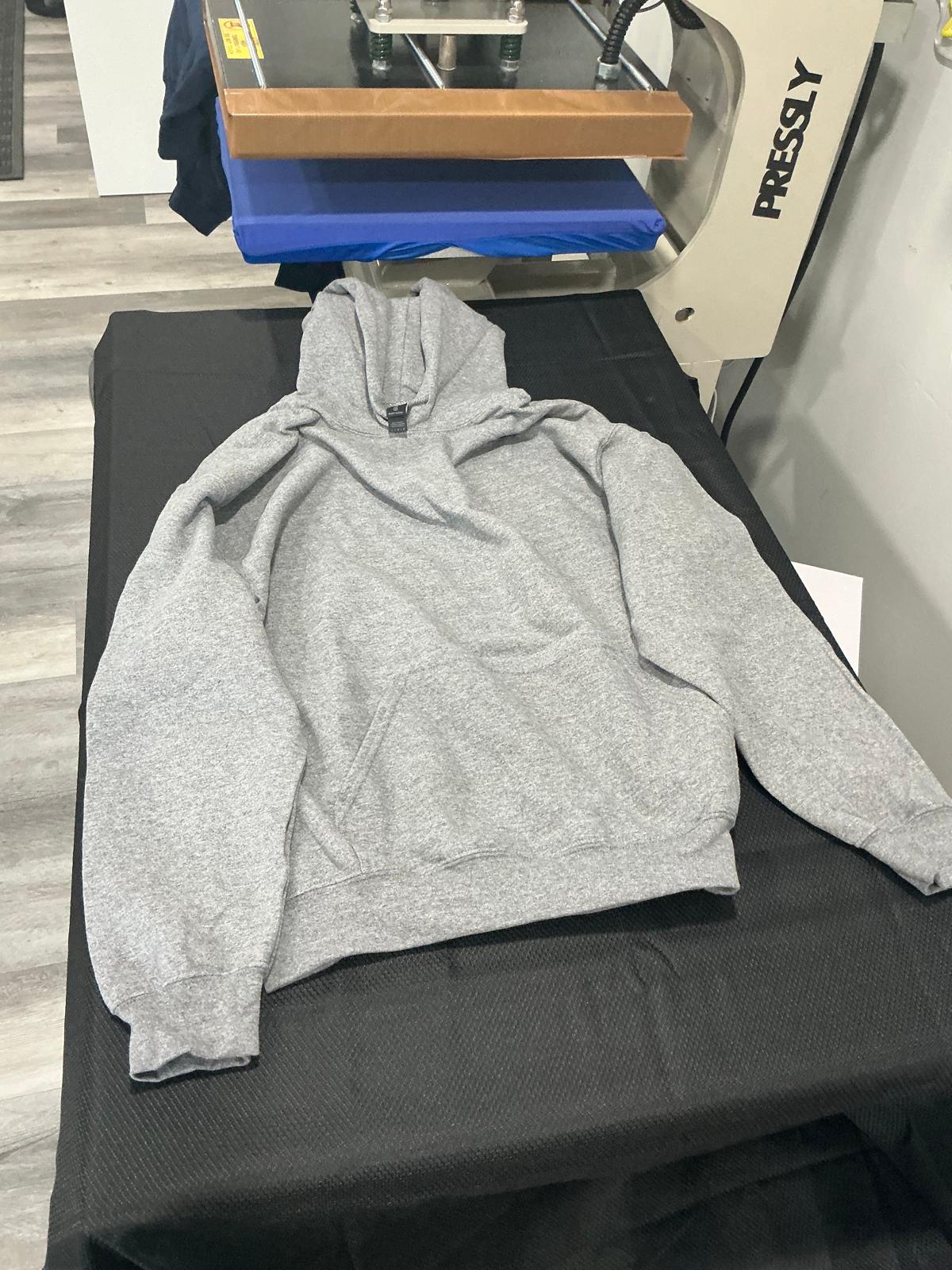 A plain heather gray heavyweight fleece pullover hoodie with a drawstring hood, laid flat and unprinted on the black work surface beside the heat press