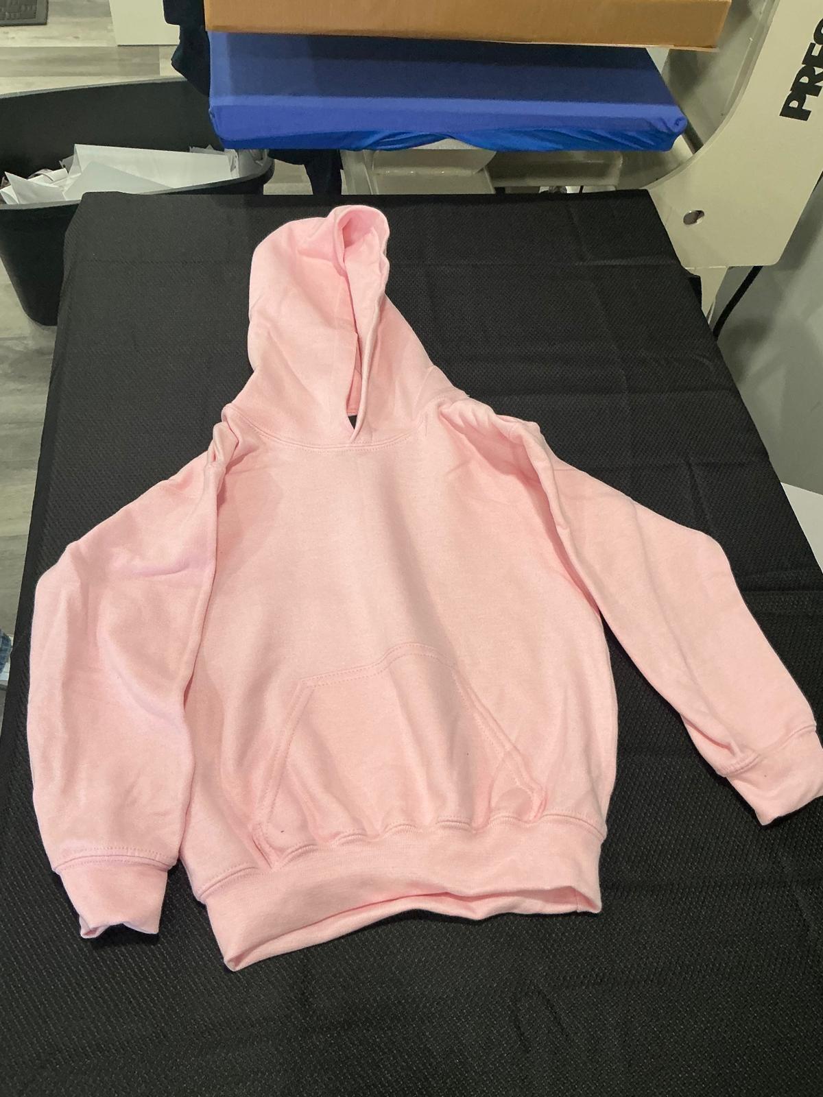 Una sudadera lisa de polar en rosa claro, para niño, con capucha y bolsillo canguro, extendida y sin estampar sobre la superficie negra de trabajo junto a la prensa de calor