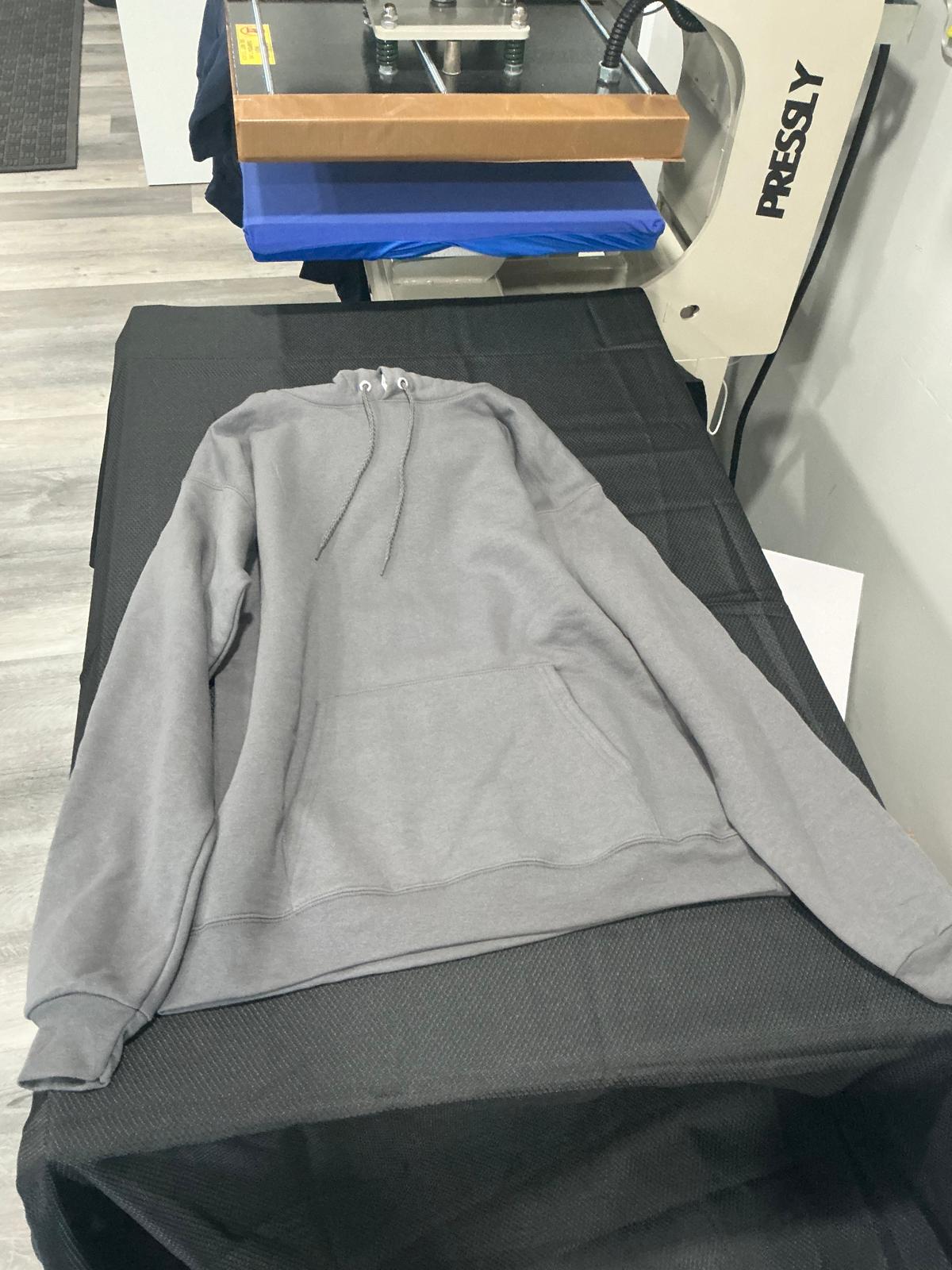 A plain gray fleece pullover hoodie with a drawstring hood and a kangaroo pocket, laid flat and unprinted on the black work surface beside the heat press