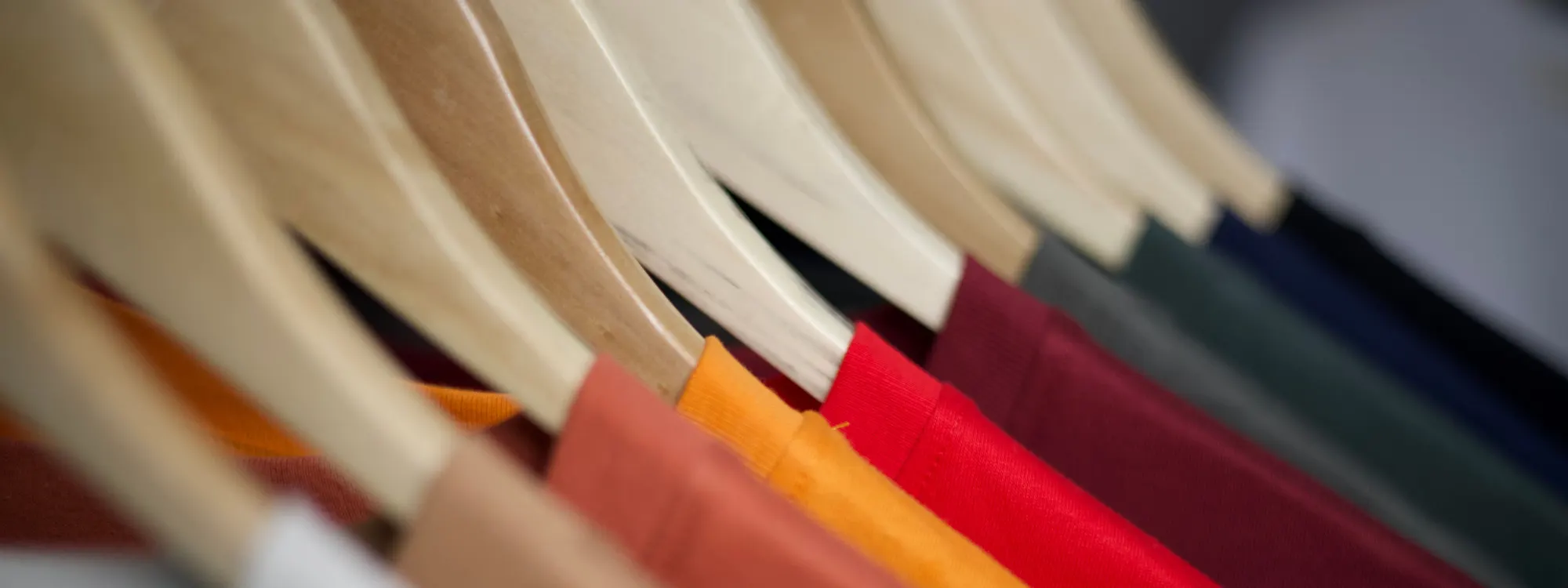 Plain, unprinted shirts in orange, coral, red, burgundy and teal hanging shoulder to shoulder on wooden hangers