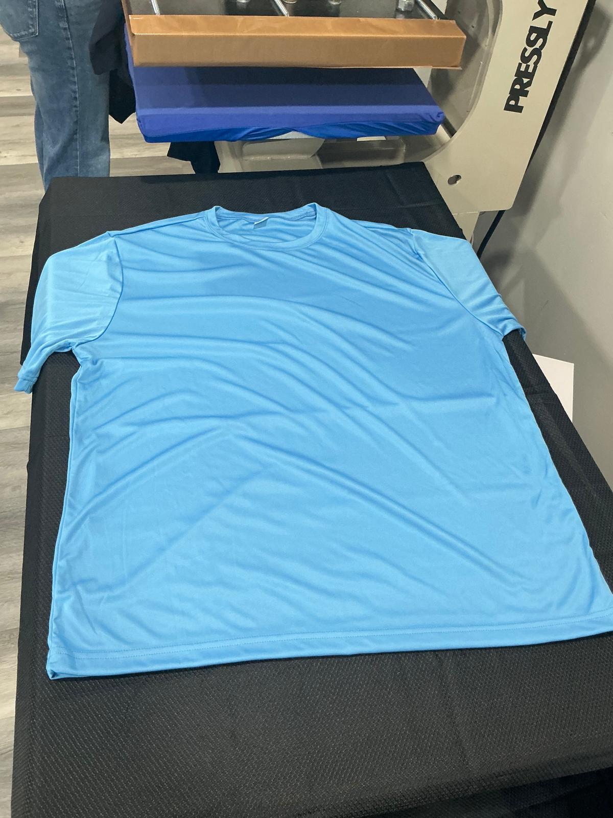 A plain light blue short-sleeve performance t-shirt laid flat and unprinted on the black work surface beside the heat press